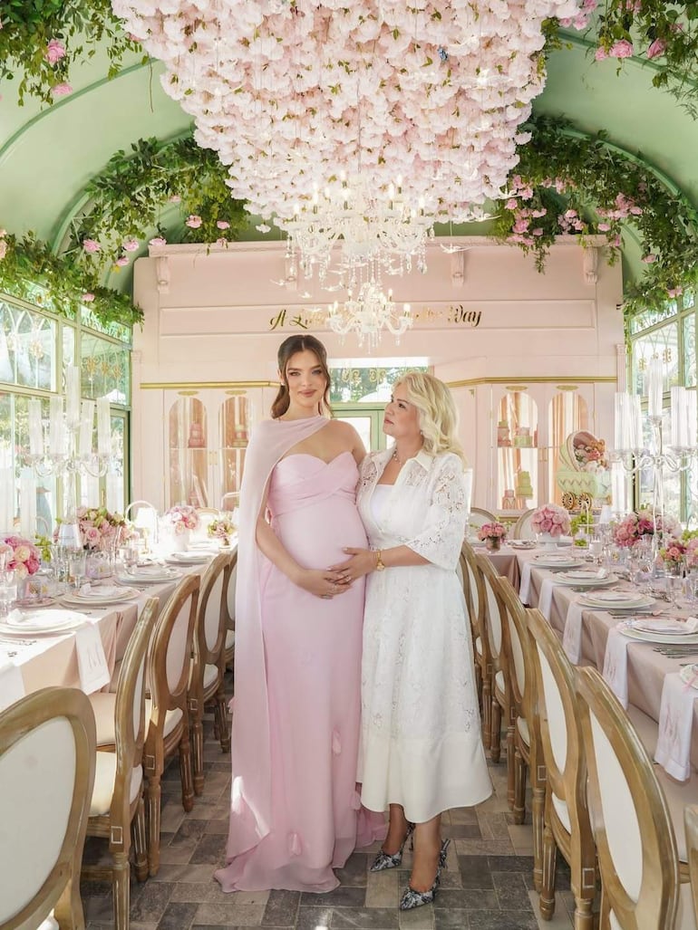 Nadia Ferreira en vestido rosa, sonriente y embarazada, conversa con mujer vestida de blanco en salón decorado con flores.