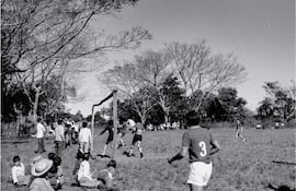 Grupo de niños sentados en el césped, mirando a los jugadores en acción en un partido sin equipo identificable.