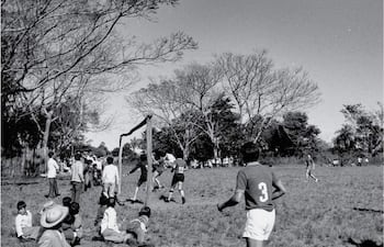 Grupo de niños sentados en el césped, mirando a los jugadores en acción en un partido sin equipo identificable.
