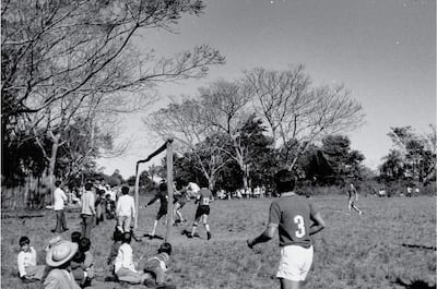 Grupo de niños sentados en el césped, mirando a los jugadores en acción en un partido sin equipo identificable.