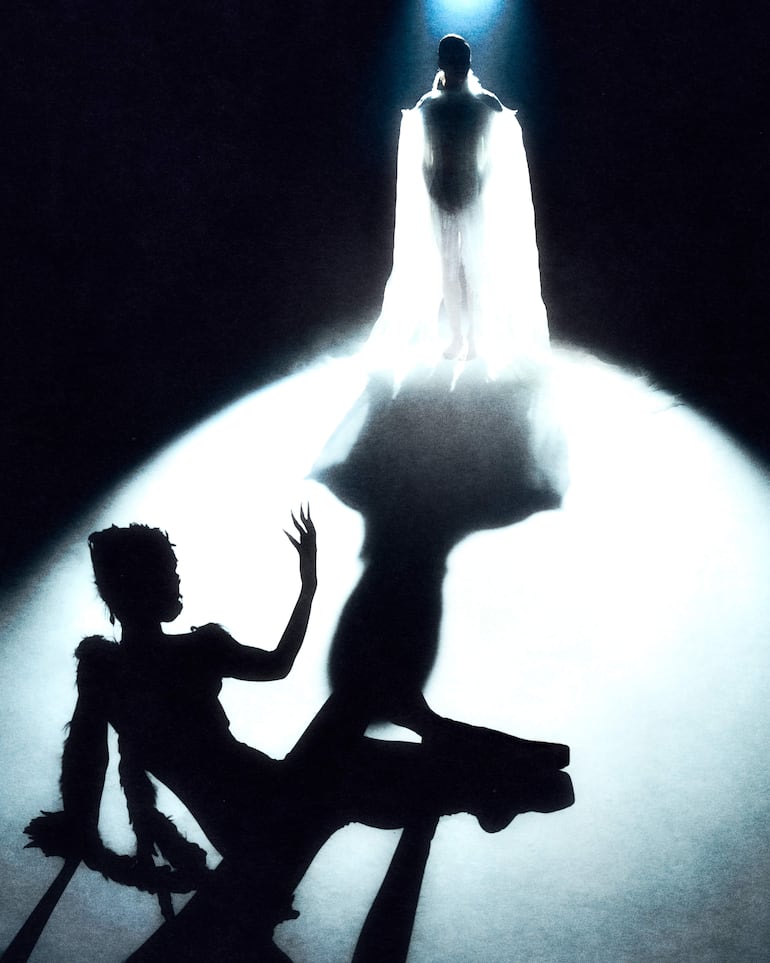 Dos figuras en escena: la principal de pie con vestido etéreo, y otra sentada gesticulando, en un entorno oscuro y dramático.