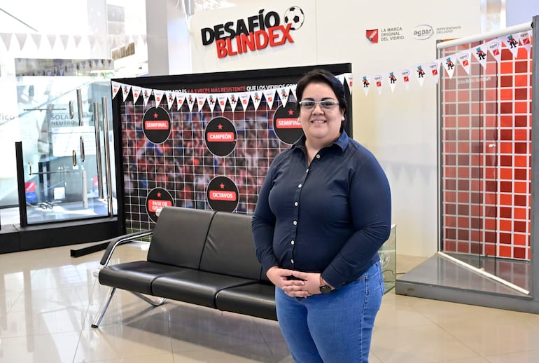 Mujer sonriente con camisa oscura y jeans, posando frente a un fondo decorativo del evento 'Desafío Blindex'.