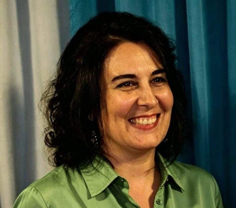 Mujer con cabello rizado y oscuro, sonriendo con camisa verde en un entorno interior formal.