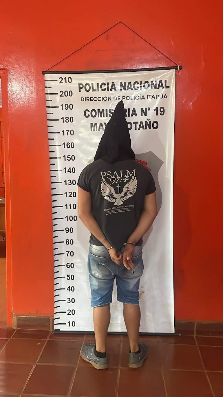 Hombre con capucha negra y camiseta con estampado, esposado en una comisaría con fondo rojo y cartel de Policía Nacional.