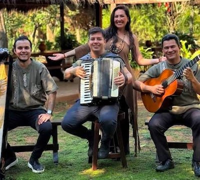 Músicos: tres hombres tocando acordeón y guitarra, y una mujer de pie con gestos festivos, rodeados de vegetación.