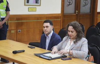Hernán Rivas, con camisa de cuadros y saco azul, y Alba Zaracho, en blazer de cuadros, revisan documentos en un ambiente judicial.