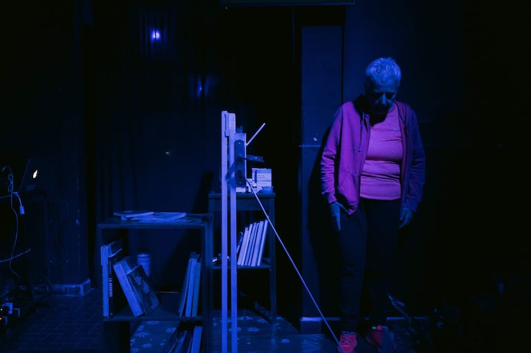 Hombre de cabello canoso y sudadera rosa, con manos inactivas, en un entorno con luz azul y estantería de libros.