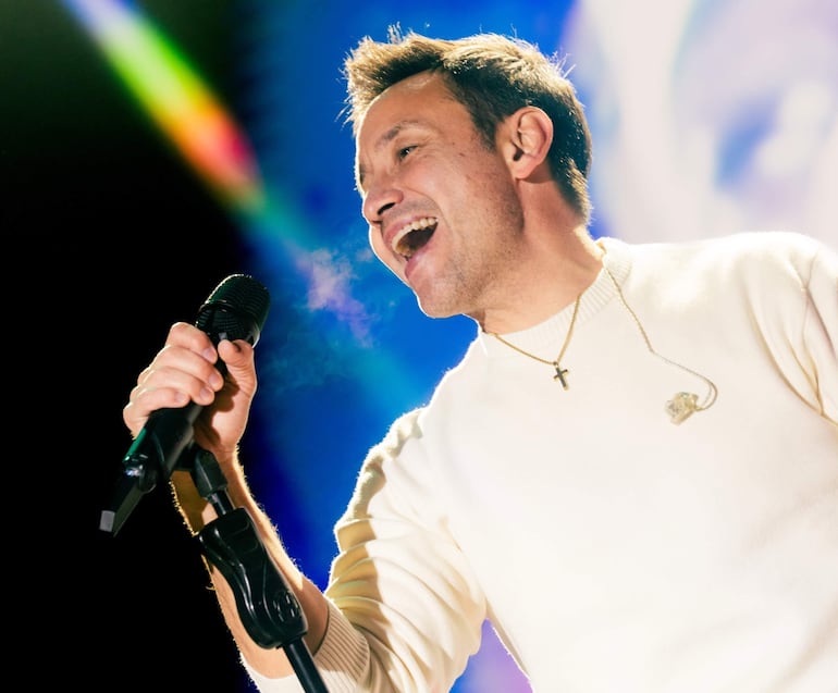 Luciano Pereyra, con suéter blanco y collar, canta con alegría en un escenario iluminado durante su actuación.