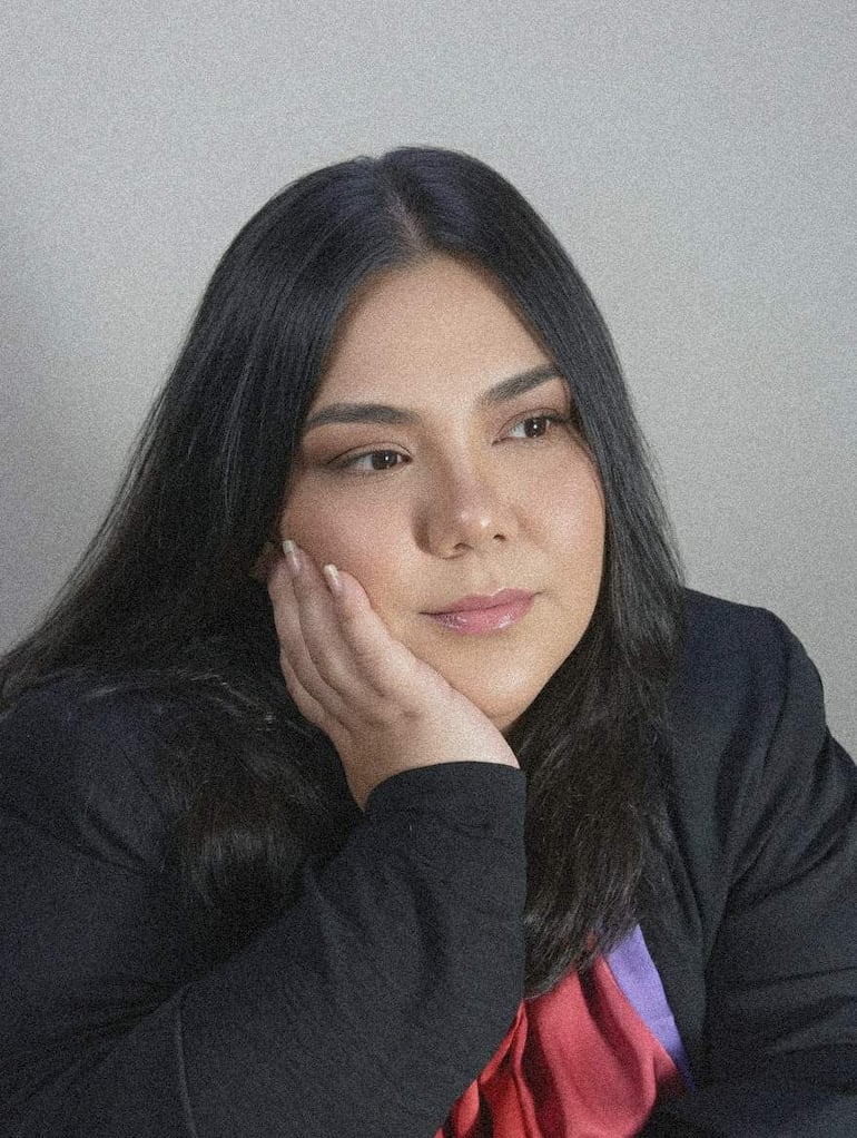 Mujer con cabello oscuro, chaqueta oscura y blusa colorida, en actitud pensativa con mano en el rostro.
