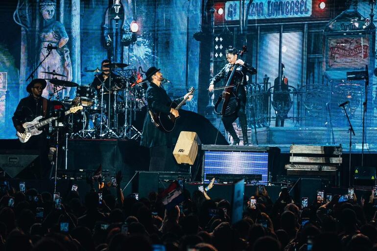 Ricardo Arjona sostiene una guitarra en el escenario, rodeado de músicos. El público graba, con un ambiente festivo y colorido.