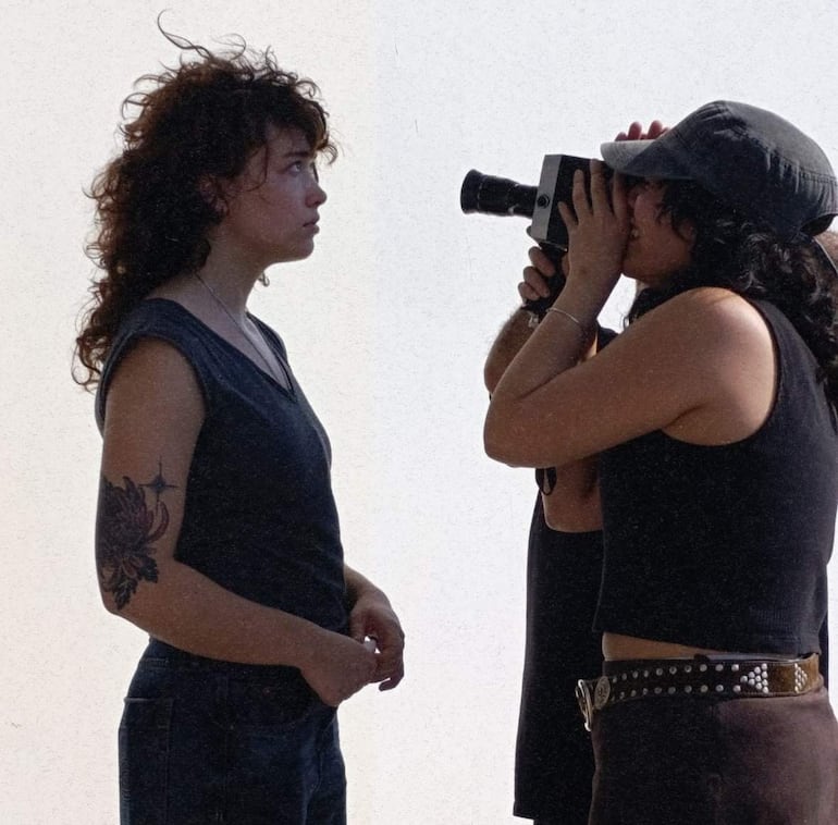Una mujer con cabello rizado y camiseta gris mira a la cámara, mientras otra, con sombrero gris, toma fotos con su cámara.