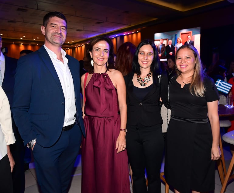 Cuatro personas sonrientes en evento social; dos hombres y dos mujeres con trajes y vestidos elegantes en ambiente decorado.