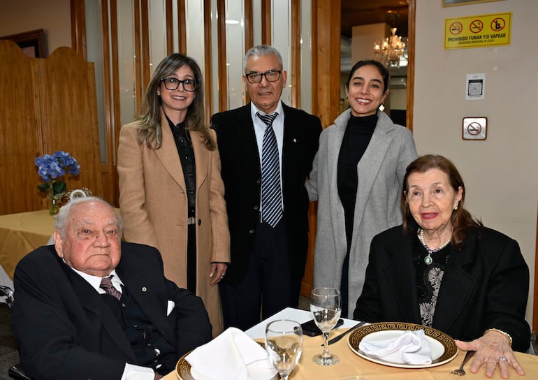 Hombre mayor en silla de ruedas y mujer mayor conversan, rodeados de tres personas más en un salón decorado con elegancia.