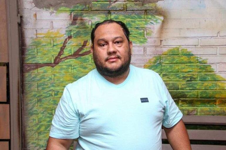 Hombre de barba y pelo rizado, con camiseta celeste, frente a un mural de árboles en un ambiente interior.