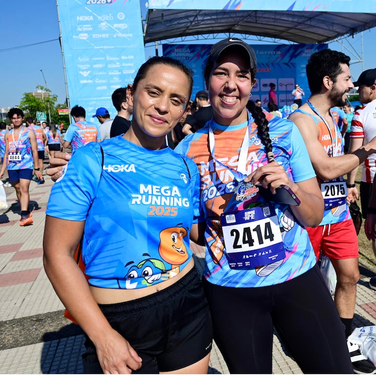 Dos mujeres con camisetas azules del evento MEGA RUNNING 2025, sonriendo y con medallas tras la carrera.