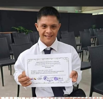 Persona con cabello corto, camisa blanca y chaleco negro sonríe mientras sostiene un certificado en sillas vacías.