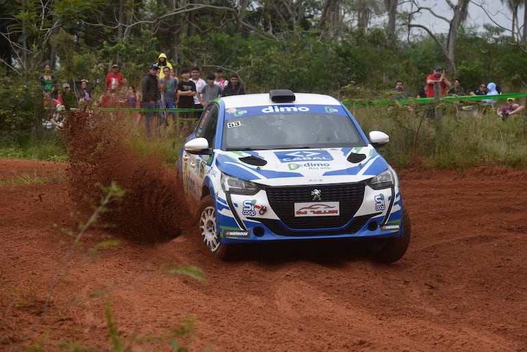 Coche Peugeot en terreno rojizo levantando polvo, rodeado de espectadores de diferentes edades, observando la carrera.