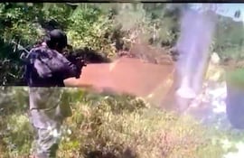 Individuo de espaldas, vestido de camuflaje, apunta un arma hacia un cuerpo de agua en zona boscosa.