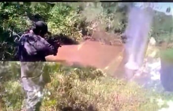Individuo de espaldas, vestido de camuflaje, apunta un arma hacia un cuerpo de agua en zona boscosa.