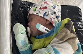 Bebé en cuna con abrigo verde y marrón, gorra blanca con autos, y chupete, en un entorno hospitalario.
