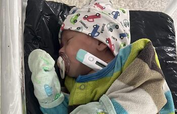 Bebé en cuna con abrigo verde y marrón, gorra blanca con autos, y chupete, en un entorno hospitalario.