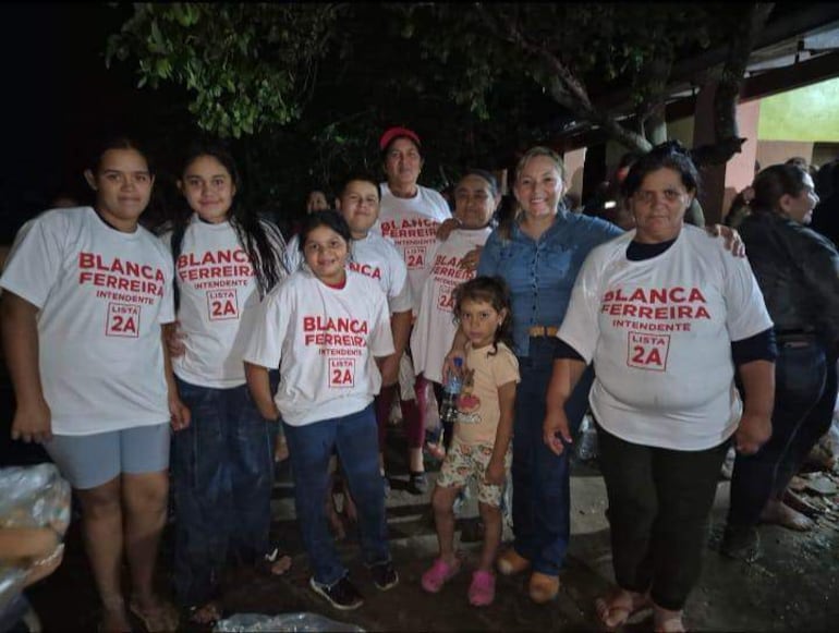Ocho personas en camisetas blancas sonríen y posan, apoyando a Blanca Ferreira en un evento nocturno con tenue iluminación.