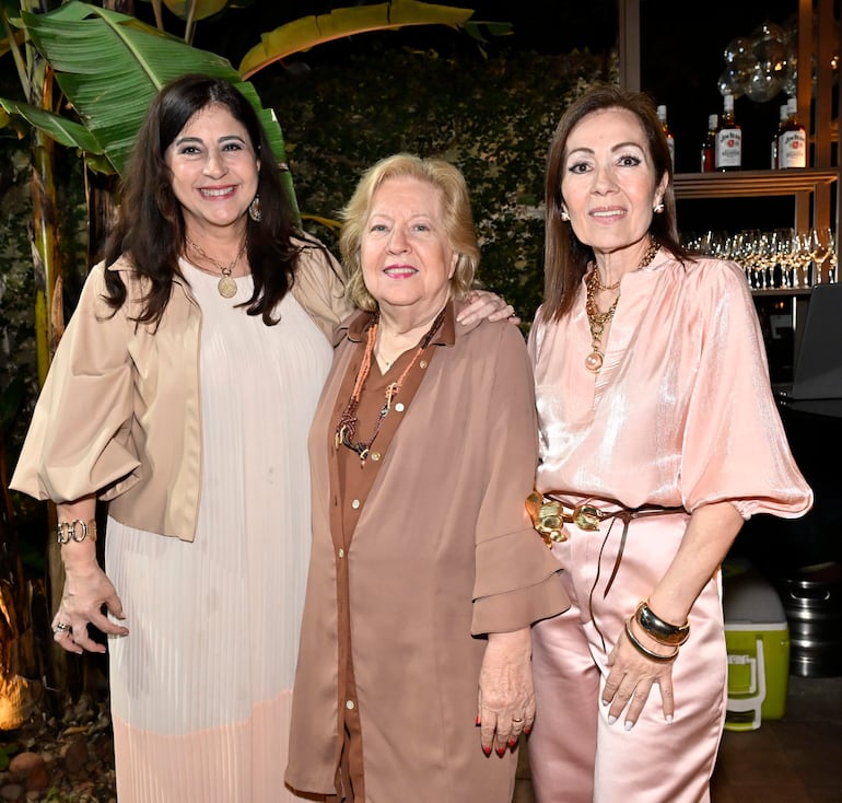 Tres mujeres sonríen en un jardín, una viste de blanco, otra de marrón y la última de rosa claro.