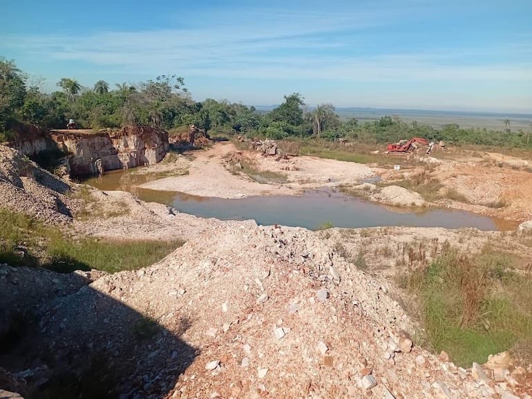 Paisaje de cantera con vehículo rojo y cuerpo de agua, rodeado de vegetación y terreno excavado.