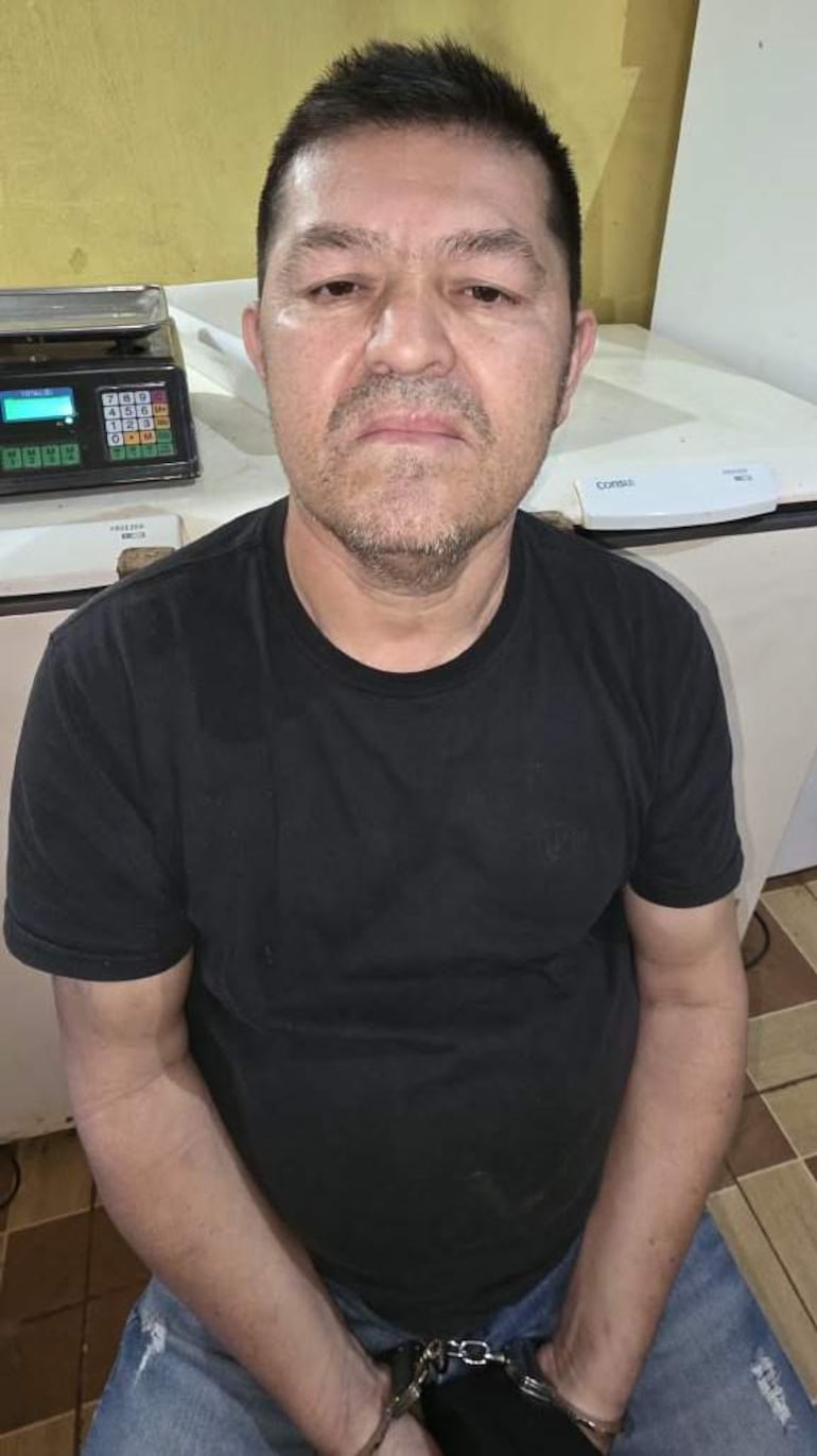 Hombre con camiseta negra y manos esposadas, expresión seria, en un entorno de escritorio con una balanza al fondo.
