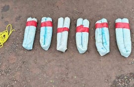 Siete paquetes de dinamita en gel sobre tierra, rodeados de cordones blanco y amarillo en un entorno exterior.