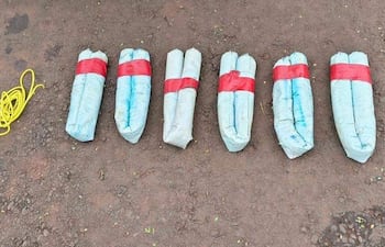 Siete paquetes de dinamita en gel sobre tierra, rodeados de cordones blanco y amarillo en un entorno exterior.
