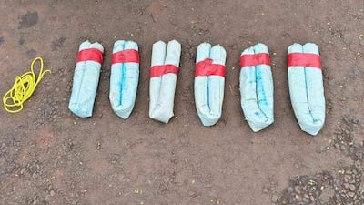 Siete paquetes de dinamita en gel sobre tierra, rodeados de cordones blanco y amarillo en un entorno exterior.