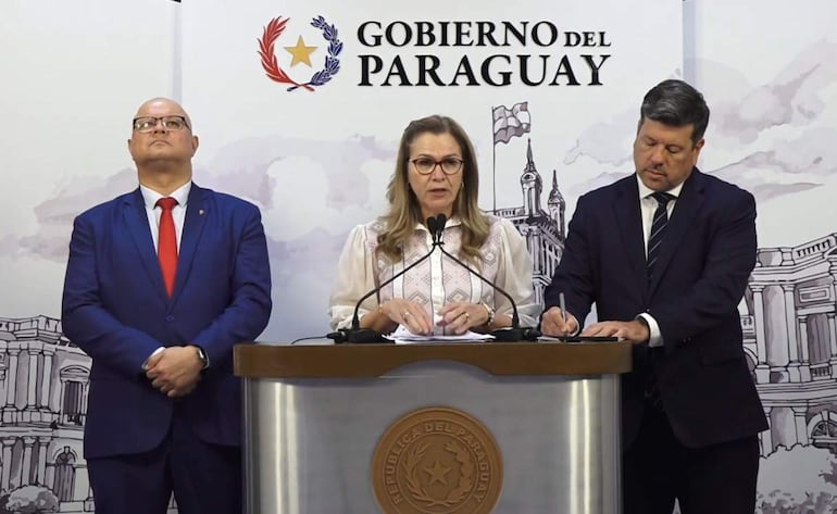 María Teresa Barán con gafas y vestimenta blanca habla en micrófono; Oscar Lovera escucha a su lado, mientras Javier Giménez toma notas.