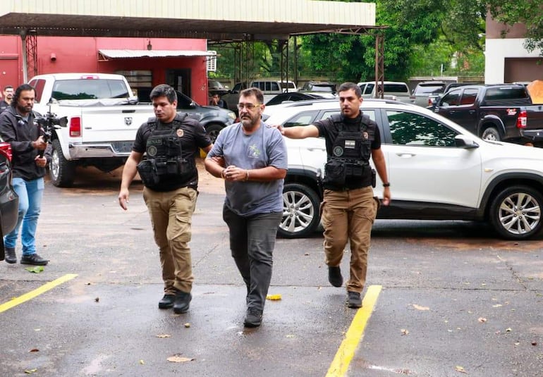 Marcos Campinha Panissa, esposado y cabizbajo, escoltado por dos agentes de seguridad en un estacionamiento.