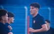 Cuatro futbolistas en camisetas oscuras entrenan en un campo, uno charla mientras otro realiza un ejercicio.