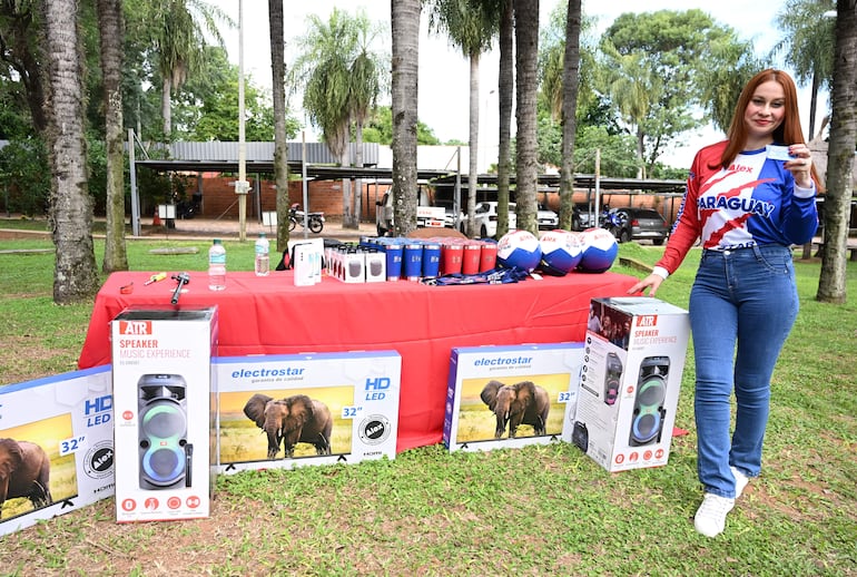 Mujer de pie con camiseta roja, blanca y azul sonríe levantando un objeto, rodeada de productos sobre mesa con mantel rojo.