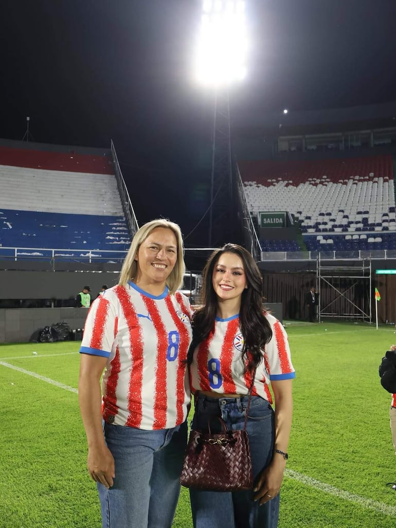 Dos mujeres sonrientes en el estadio, una con cabello rubio y la otra con cabello oscuro, ambas visten camisetas de rayas rojas y blancas.