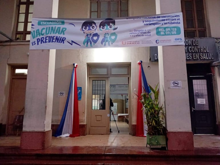Cartel de vacunación en edificio de salud, decorado con cintas de los colores de la bandera paraguaya, sin personas visibles.