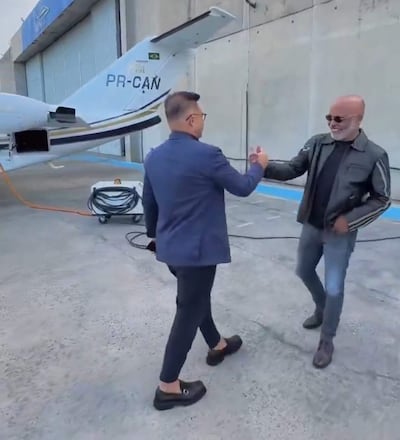 Hombre con traje azul oscuro saluda a otro con chaqueta negra y jeans en un aeropuerto cerca de un avión privado registrado como PR-CAN.
