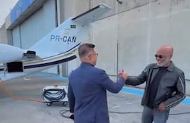 Hombre con traje azul oscuro saluda a otro con chaqueta negra y jeans en un aeropuerto cerca de un avión privado registrado como PR-CAN.