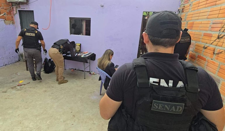 Agentes de la SENAD con vestimenta oscura revisan un escritorio mientras una mujer observa atención al teléfono móvil en un ambiente púrpura.