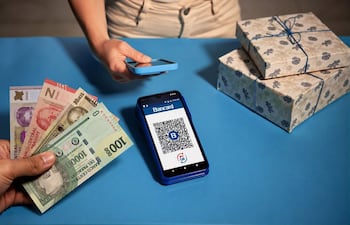 Mano con billetes de 100 y 2000 guaraníes junto a teléfono mostrando código QR de Bancard en fondo azul.