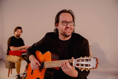 Diego Guzmán sonriente con guitarra clásica en abrigo negro, mientras un hombre en camiseta oscura lo acompaña al teclado.