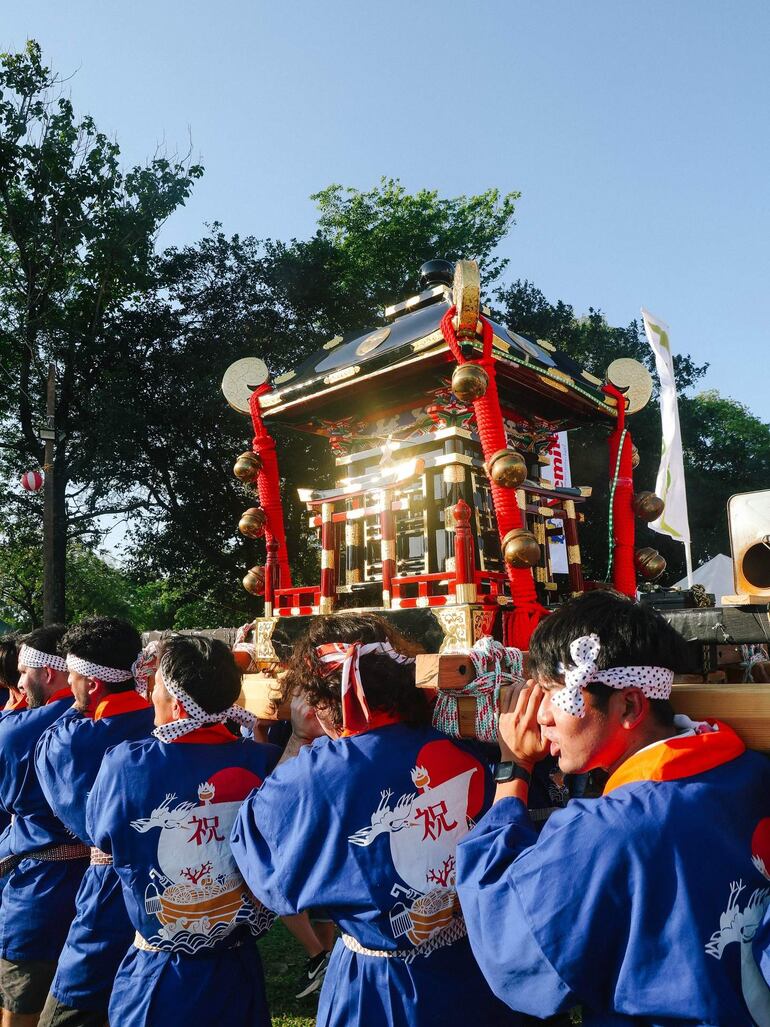 Ocho hombres con camisetas azules, pañuelos en la cabeza, levantan un mikoshi adornado en un entorno festivo al aire libre.