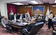 Cecilia Rodríguez habla de pie ante una mesa con documentos, mientras Juan José Galeano y otros escuchan en un ambiente formal.
