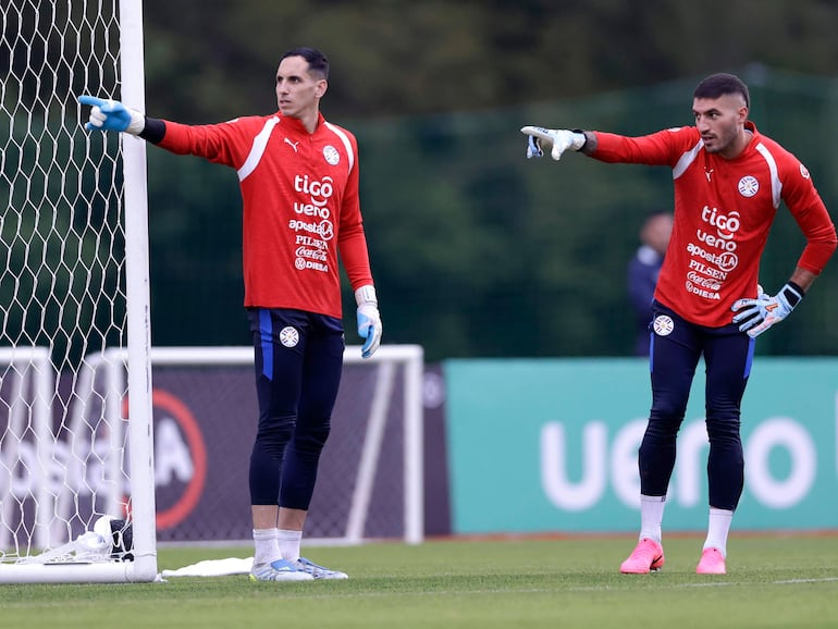 Gatito Fernández y otro portero en uniforme rojo, guantes y concentrados, entrenan en cancha de césped verde rodeada de árboles.