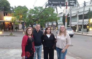 Cuatro personas conversan en la acera; mujer de cabello rizado en negro y rojo, hombre con anteojos y camisa oscura, mujer con camiseta negra y mujer rubia con camisa a rayas.