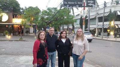 Cuatro personas conversan en la acera; mujer de cabello rizado en negro y rojo, hombre con anteojos y camisa oscura, mujer con camiseta negra y mujer rubia con camisa a rayas.