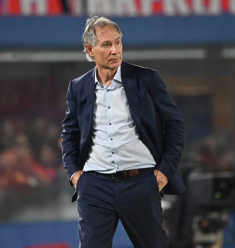 Hombre de mediana edad con cabello canoso, vestido en traje oscuro, observa el partido en un estadio con fondo de multitud.