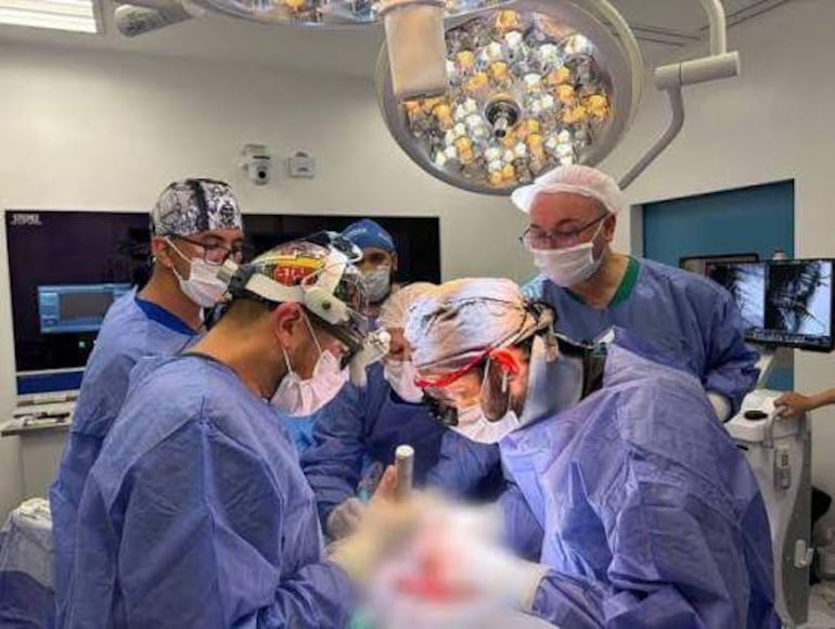 Seis profesionales de la salud en uniforme azul y mascarillas, concentrados en un procedimiento en quirófano iluminado.
